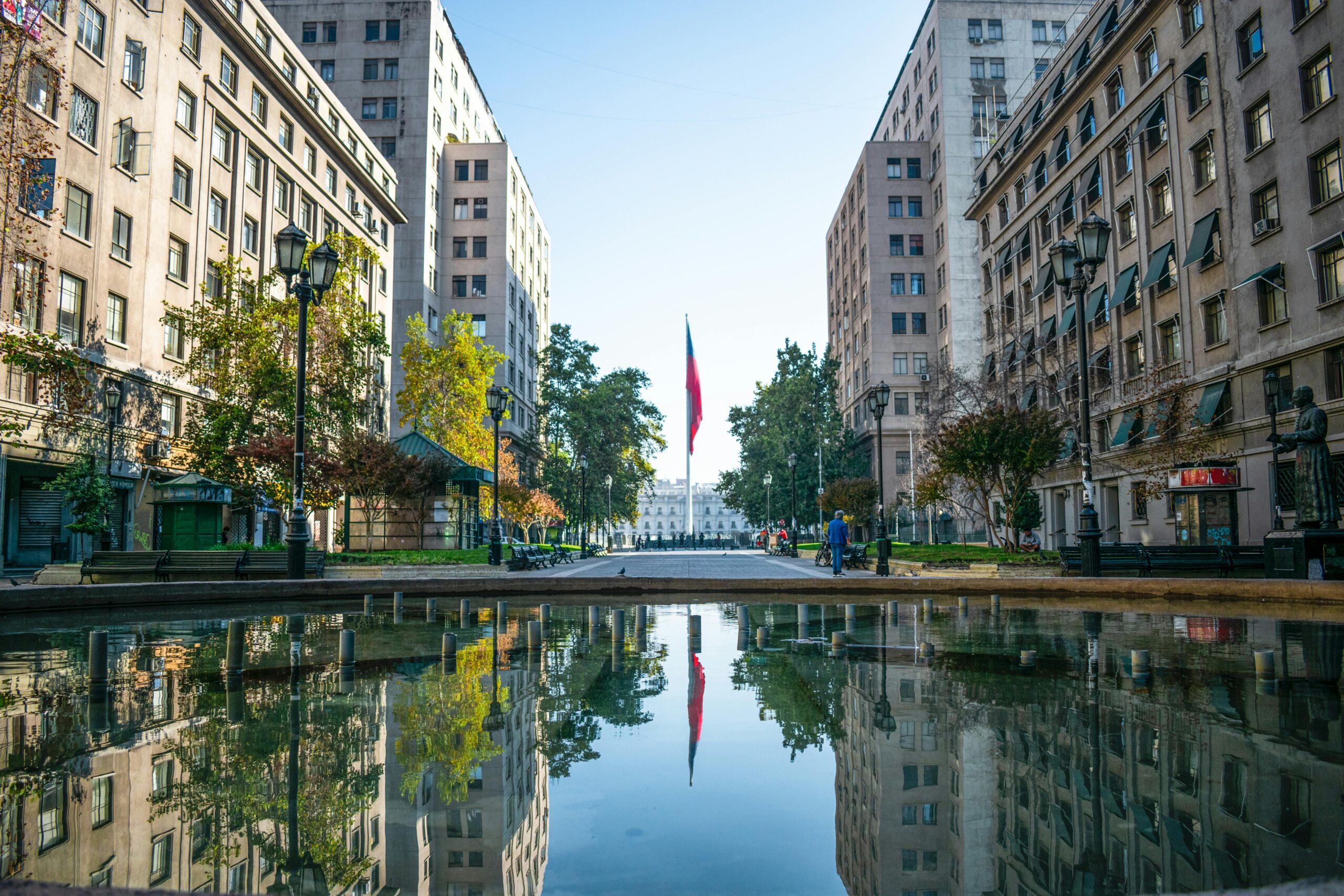 Vista urbana de Santiago Centro con edificios residenciales y bandera de Chile reflejada en una fuente de agua